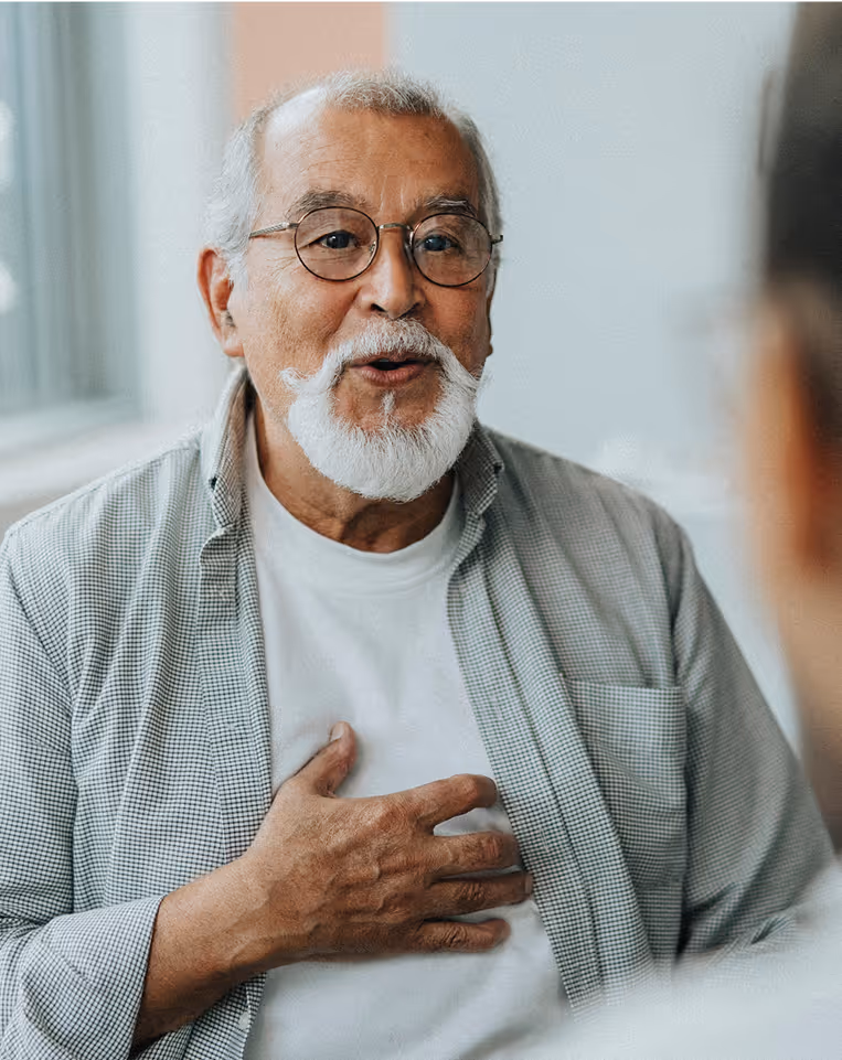 Elderly man wearing glasses and a gray shirt holding his chest while smiling.