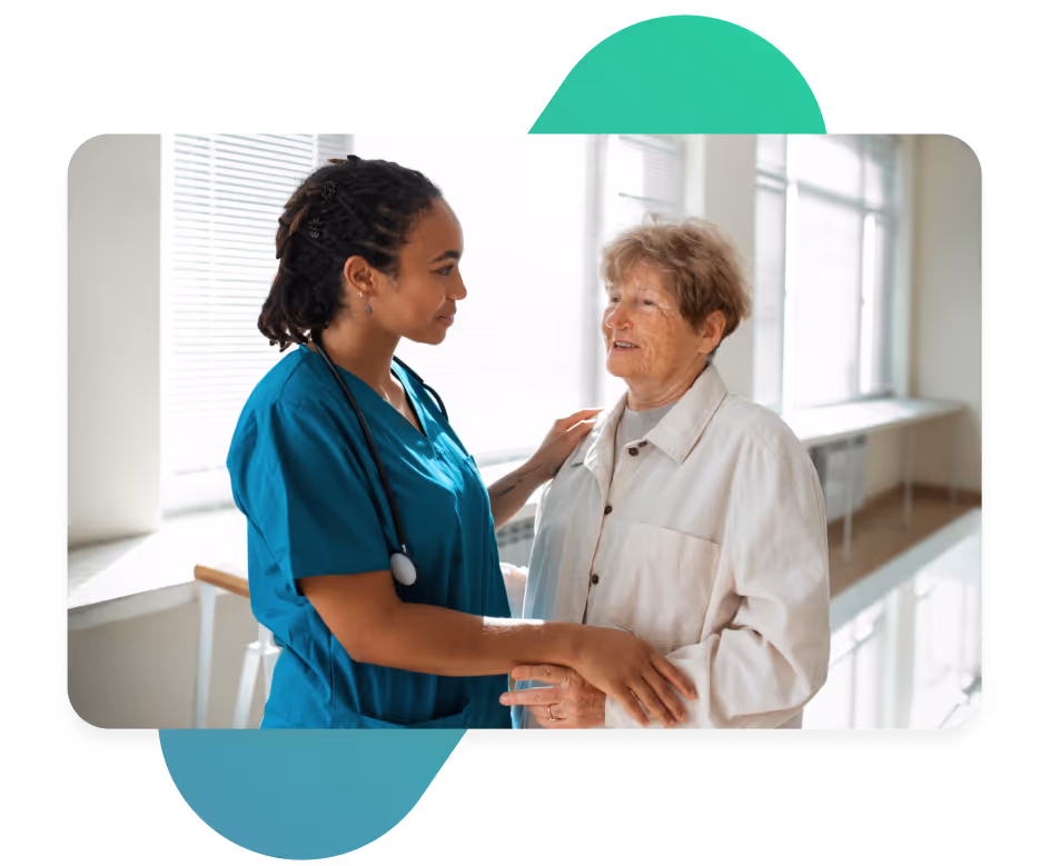 Female doctor in scrubs speaking compassionately with an elderly woman in a clinical setting.
