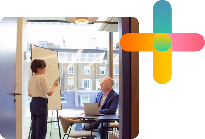 A businesswoman presenting on a flipchart to a businessman with a laptop in a modern office meeting room, next to a colorful abstract plus-shaped graphic.